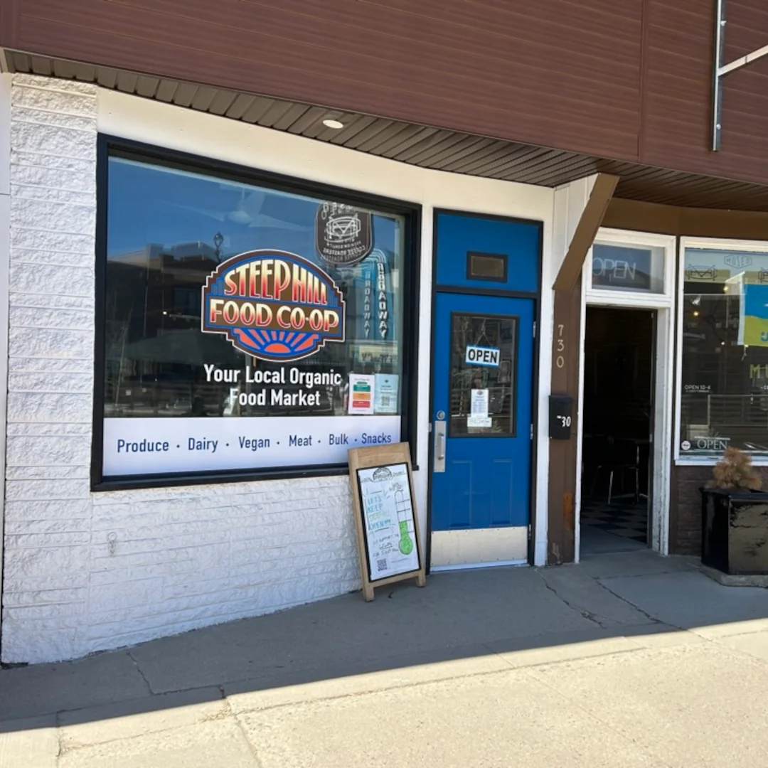 The store front of Steep Hill Food Co-op with largely visible vinyl window sign of the Steep Hill logo with text that reads, Your Local Organic Food Market.