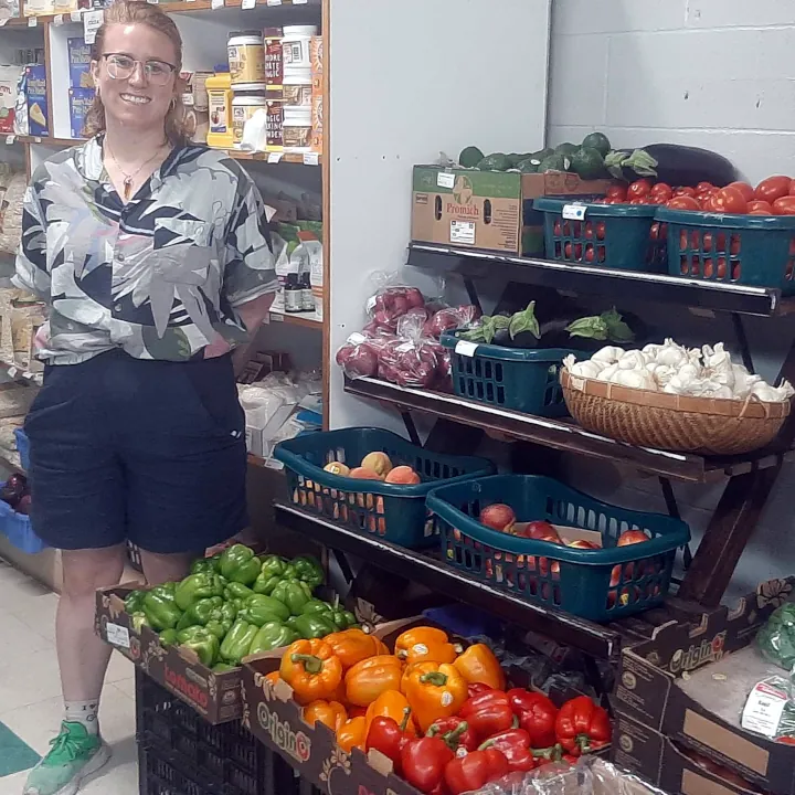 Steep Hill Food Co-op store manager Amielle stands next to a dark brown wooden shelves displaying fresh and vibrant coloured produce.