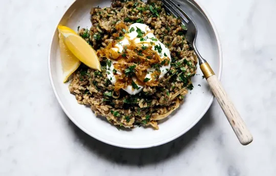 A fork leans on the side of the plate full of brown lentils garnished with two slices of lemons, sour cream and spinach.