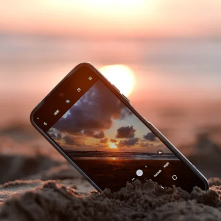 A mobile phone pushed into sand on a beach that is capturing the beauty of the summer sunset off in the distance.