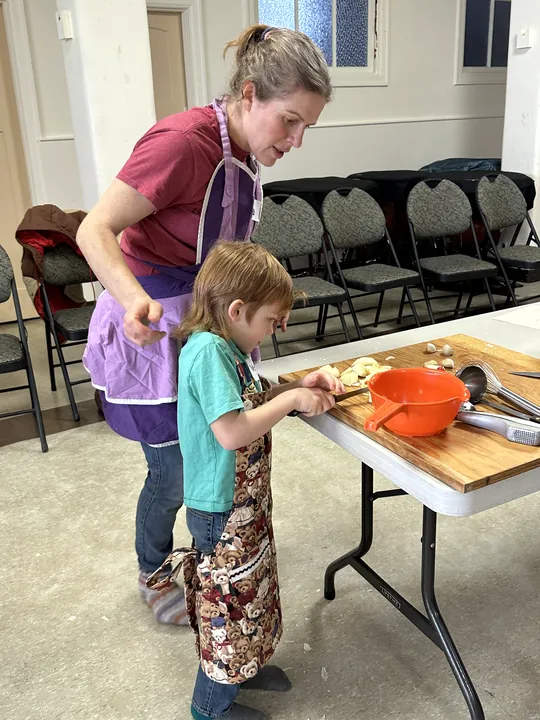 A woman and child standing in front of a table as the woman instructs the child how to chop the apples.