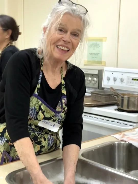 A woman with both her hands in a sink washing dishes as she smiles towards the camera.
