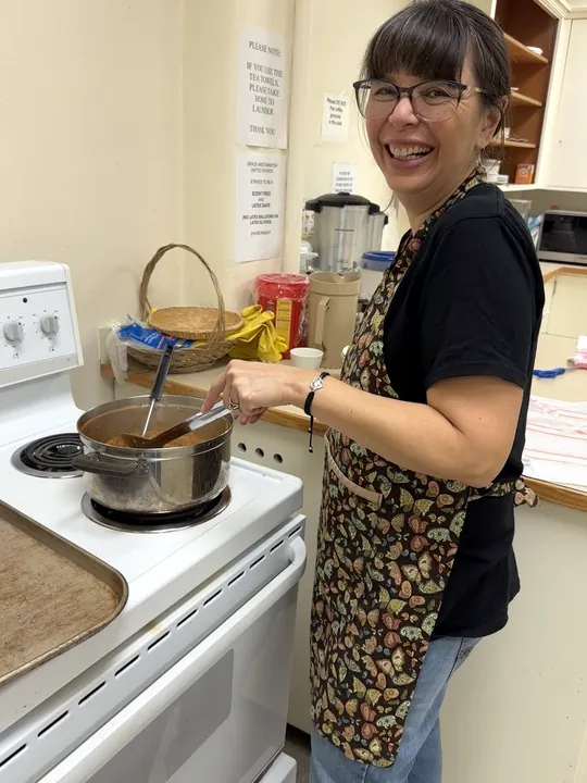 A woman standing in front of a stove with a big smile as she stirs the Mulligatawny soup.