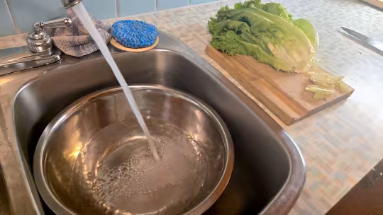 A stainless steel bowl sitting in a sink being filled up with cold water. Close by on the counter is a wooden cutting board with lettuce.