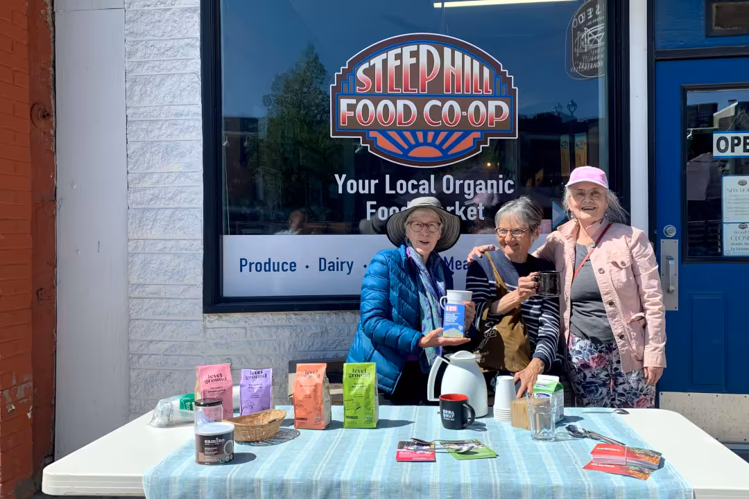 Three ladies standing in front of Steep Hill by a table that is displaying fair trade products for Fair Trade Day.