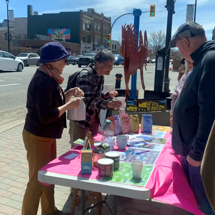 A group of people are talking next the Steep Hill Food Co-op table setup on the sidewalk facing Broadway Avenue. The table has a variety of goods on display.