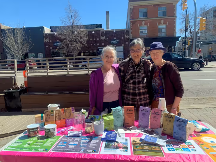 Three Steep Hill members are posing together in front of Steep Hill's table displaying a variety of goods for sale for the Fairtrade Day.
