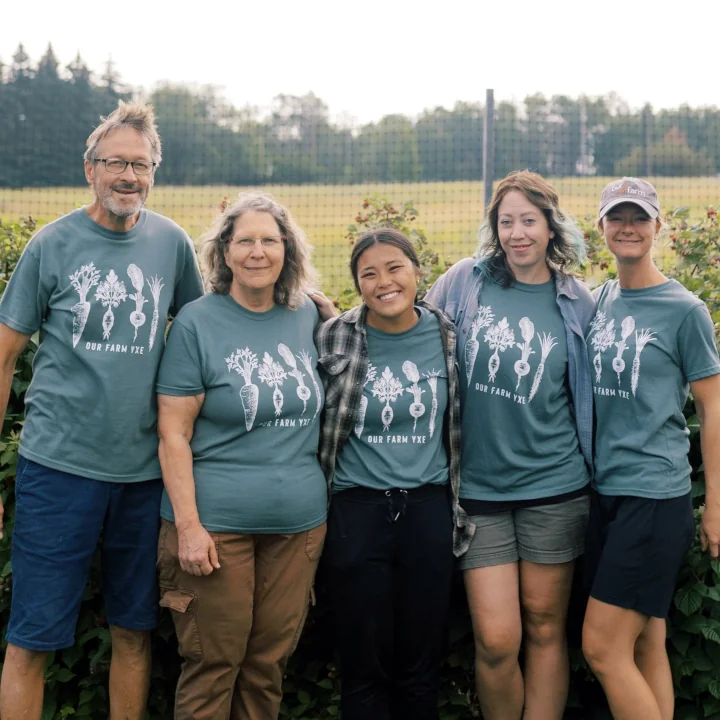 Five staff members of Our Farm YXE standing side by side wearing matching t-shirts happily posing against a farm field.