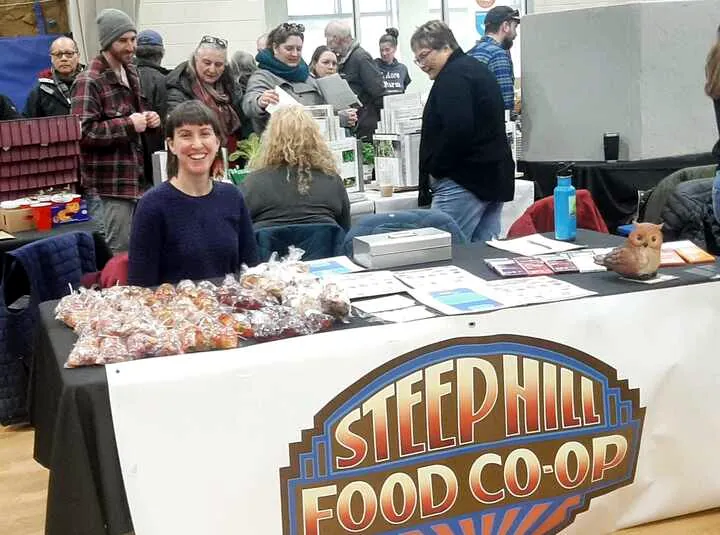 A large table draped with a black table cloth and a white background sign hangs over the front of the table with the Steep Hill Food Co-op logo. A volunteer sits behind the table full with goodies.