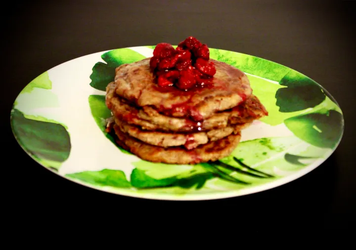 A plate with a green leaf pattern sits on a dark coloured counter with four whole grain pecan pancakes in the centre topped with raspberries, avocado oil margarine and maple syrup.