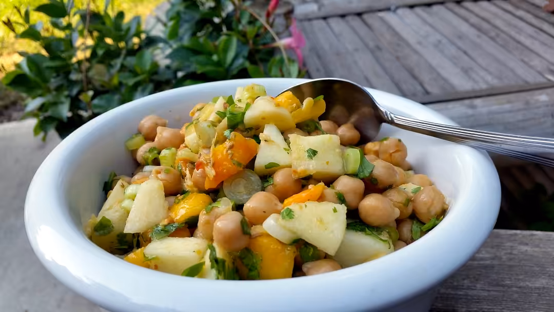 A white bowl full of a colourful Chickpea Fruit Salad and a silver spoon placed on top.