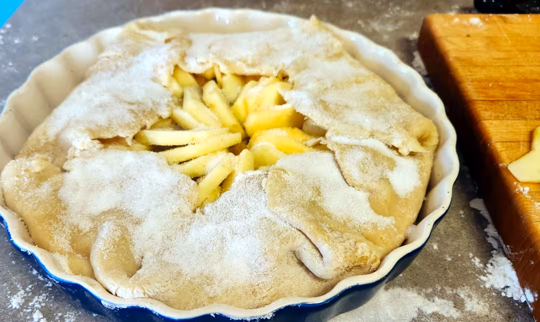 A blue and white pie plate containing fresh pastry batter covered in white sugar that has been folded over towards the center leaving the center of the tart open exposing the apples.
