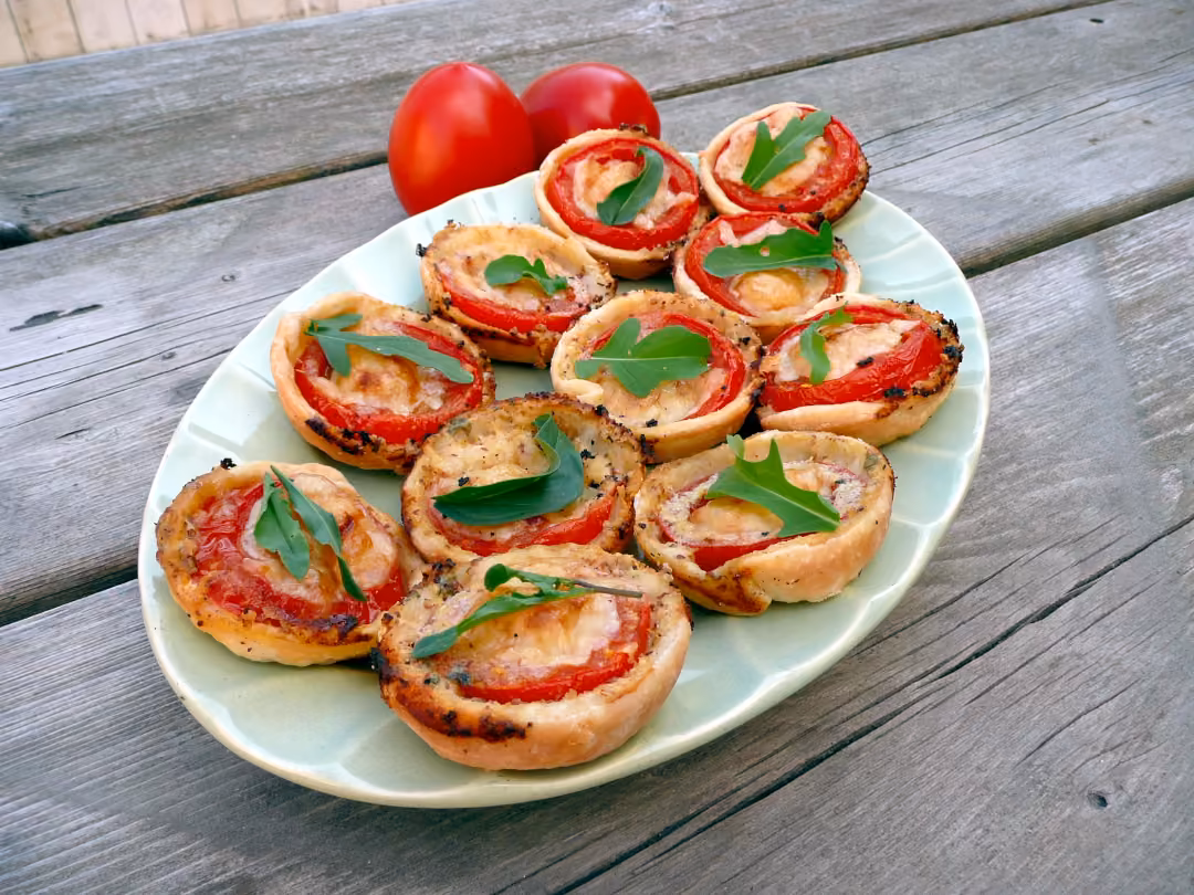 On a rustic wooden table sits an oval platter full of fresh tomato tarts topped with arugula. Off to one side of the platter is two red ripened tomatoes.