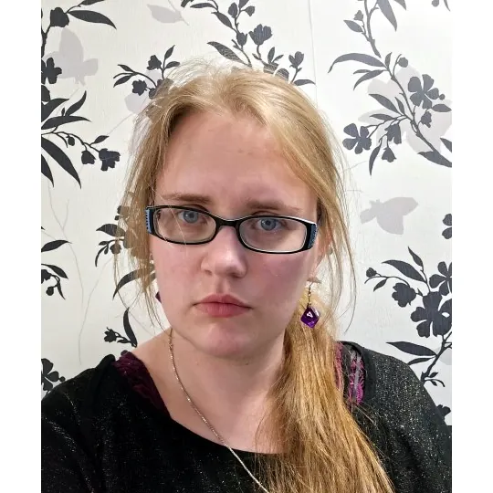 A portrait of staff member Sol gazing into the camera with blue eyes, purple earrings and rectangle shaped glasses.