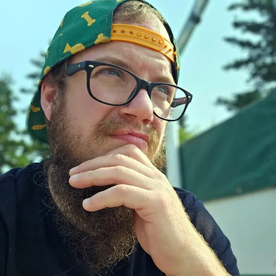 Staff member Stephan leans to one side with hand on chin looking into the distance while wearing a baseball cap backwards and black framed glasses.